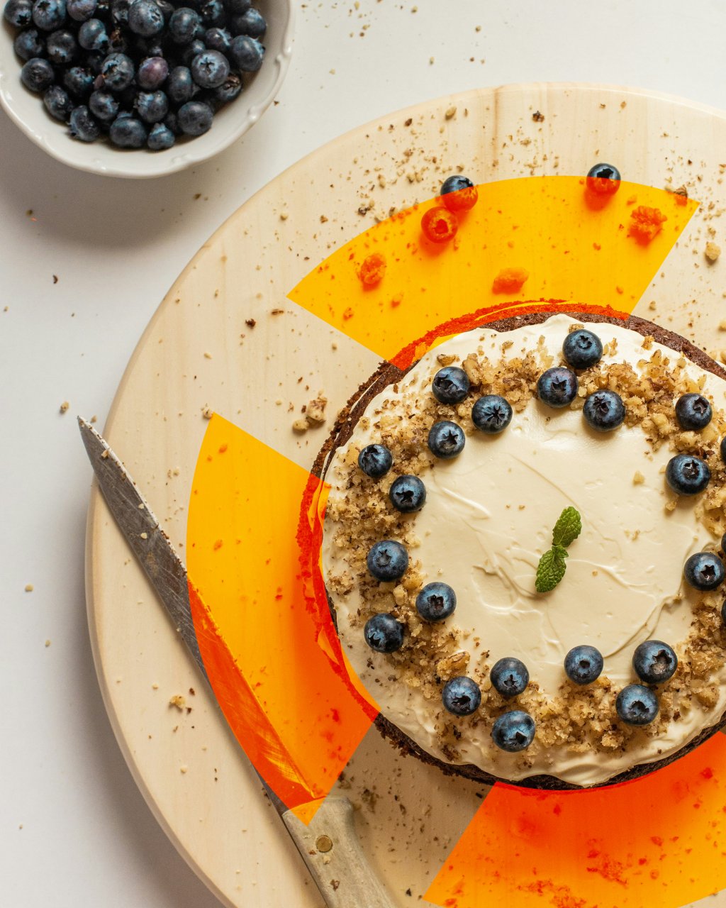 Kuchen mit Frischkäse-Frosting, Blaubeeren, Nusskrümeln und Minzblatt auf Holzbrett, Messer und Schale mit Blaubeeren