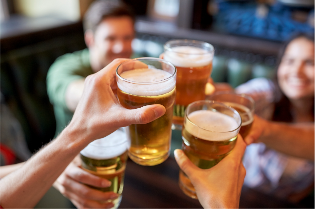 Mehrere Hände stoßen mit vollen Biergläsern an, im Hintergrund verschwommene Personen.