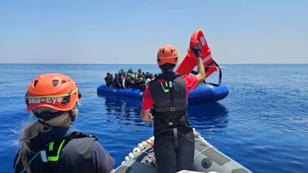 Rettungskräfte mit orangen Helmen auf Boot, eine hält Schwimmweste vor überfülltem blauen Schlauchboot auf dem Meer
