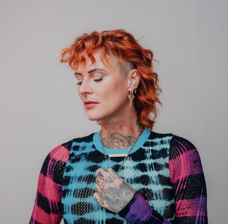 Foto von YAENNIVER (Jennifer Rostock), sie hat die Augen geschlossen, ist im Halbprofil zu sehen, Haare rot gefärbt, schulterlang, großflächige Tattoos an Hals und Handrücken, auffälliger Schmuck an Nase und Ohr, farbkontrastige Oberkörperbekleidung