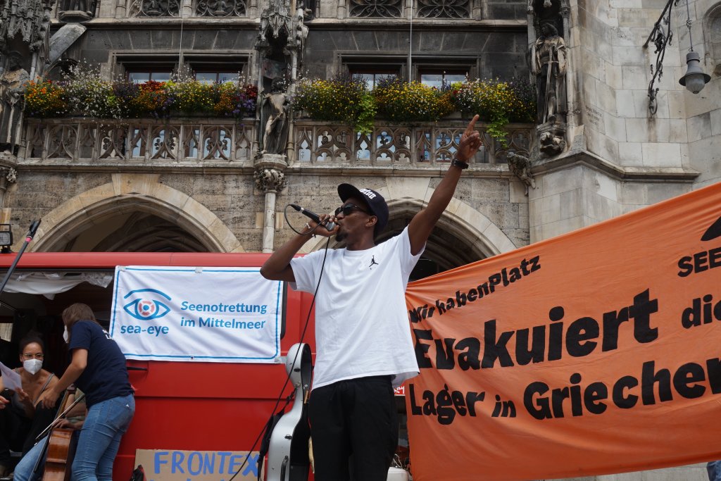 Mann mit Mikrofon und erhobenem Arm vor historischem Gebäude mit Blumenbalkon und orangefarbener Protestfahne